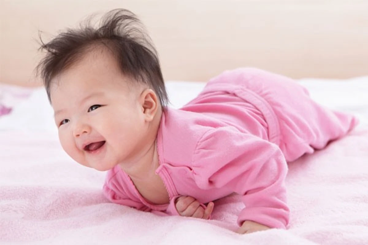 Trẻ sơ sinh mặc bộ đồ màu hồng đang nằm sấp ngẩng cao đầu vui vẻ trên đệm