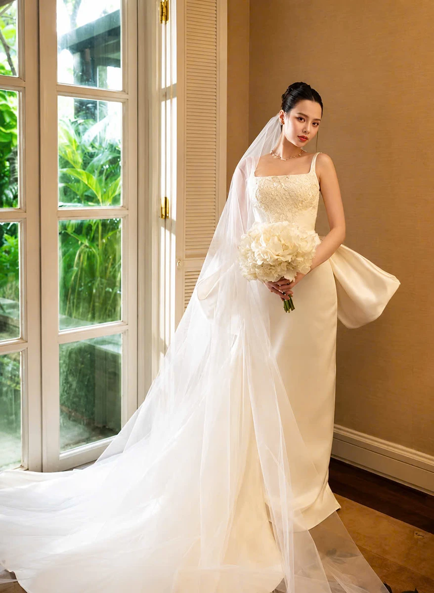 Cô dâu đứng nghiêng người bên cửa sổ, hai tay ôm bó hoa cưới và nhìn về máy ảnh, khoe dáng váy cưới cùng tà voan dài