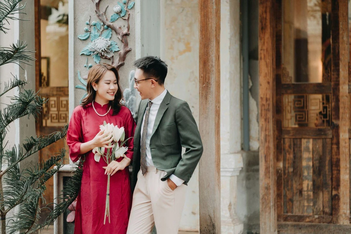 Cô dâu chú rể đứng cạnh nhau trước hiên nhà, cùng nhìn nhau mỉm cười trong tư thế chụp ảnh cưới đời thường