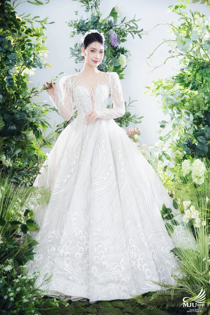 Cô dâu mỉm cười rạng rỡ trong mẫu váy cưới công chúa bồng bềnh thêu họa tiết sóng nước lấp lánh, đứng kiêu sa giữa bối cảnh sân khấu ngập tràn sắc xanh tự nhiên