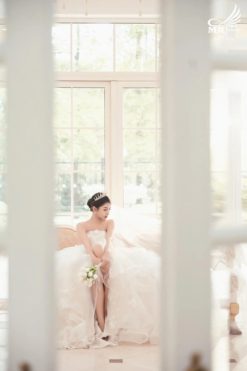 Cô dâu đội vương miện kiêu sa ngồi trên ghế sofa, diện mẫu váy cưới cúp ngực bồng bềnh lãng mạn kết hợp cùng bó hoa cưới màu trắng cầm tay dịu dàng