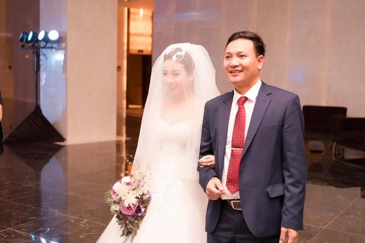 Cô dâu khoác khăn voan cầm bó hoa cưới tím trắng, sánh bước cùng người thân trong khoảnh khắc tiến vào lễ đường