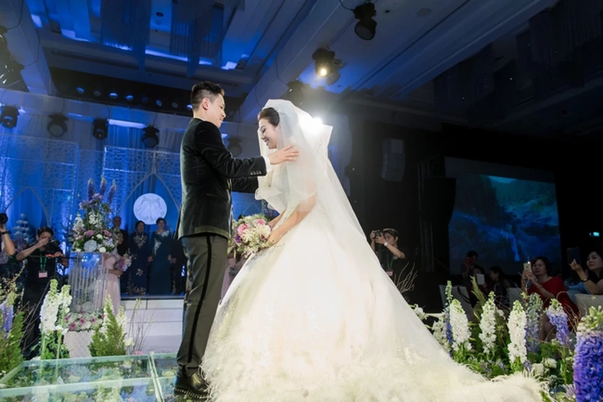 Chú rể chỉnh lại khăn voan cho cô dâu trên sân khấu lễ cưới, giữa không gian trang trí hoa tươi và lối đi kính