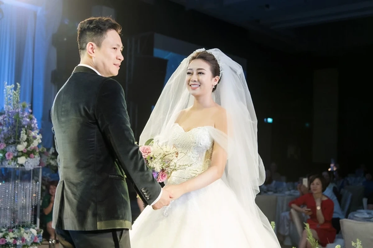 Cô dâu chú rể nắm tay nhau mỉm cười hạnh phúc trên sân khấu tiệc cưới với váy cưới trắng và vest đen lịch lãm