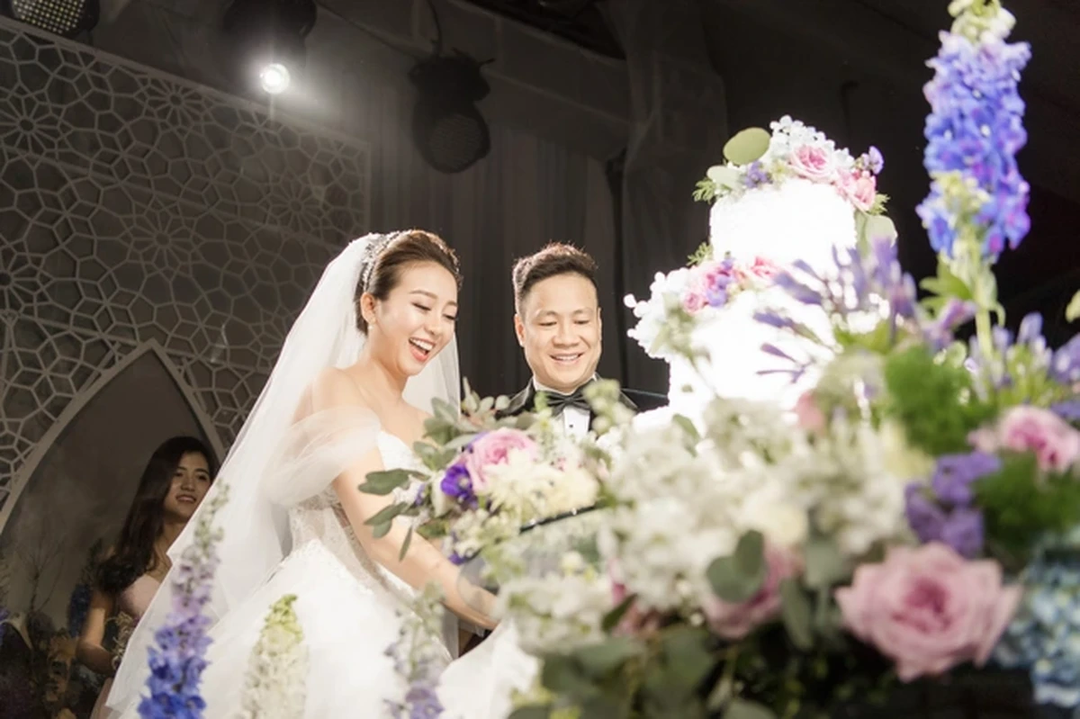 Cô dâu chú rể cùng cắt bánh cưới nhiều tầng được trang trí hoa tươi tone tím, trắng