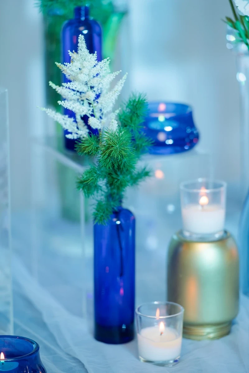 Bình thủy tinh xanh kết hợp hoa trắng, lá xanh và nến trang trí, mang phong cách decor cưới nhẹ nhàng, hiện đại