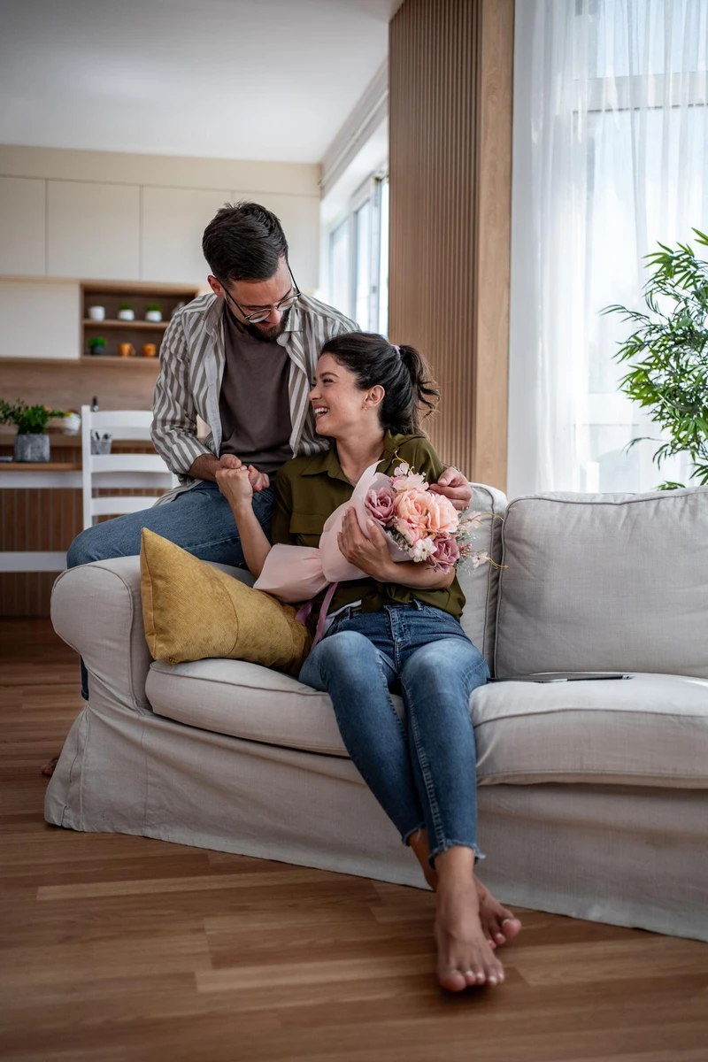 Người đàn ông tặng bó hoa tươi thắm cho bạn gái trên ghế sofa phòng khách, tạo nên khoảnh khắc bất ngờ và lãng mạn trong tình yêu