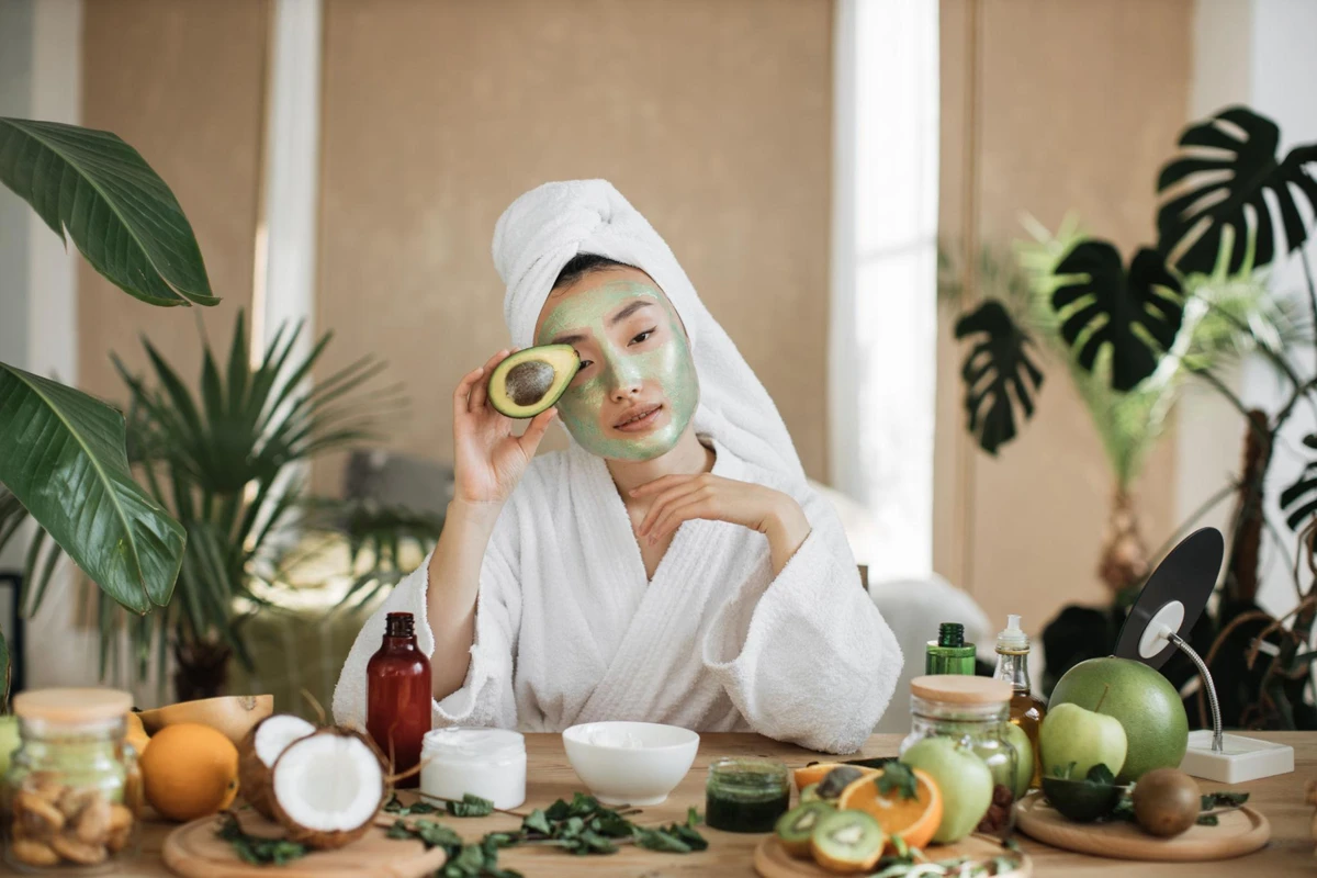 Cô gái quấn khăn tắm màu trắng đang thoa mặt nạ bơ dưỡng da, một tay cầm nửa quả bơ tươi trước bàn trang điểm chứa nhiều loại trái cây giúp chăm sóc da mặt
