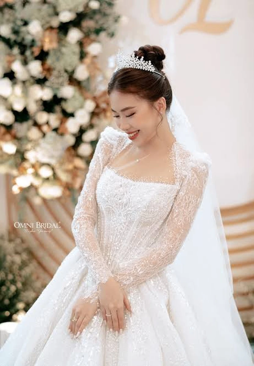 Cô dâu diện váy cưới trắng tay dài đính sequin lấp lánh, kết hợp vương miện và khăn voan, mỉm cười dịu dàng