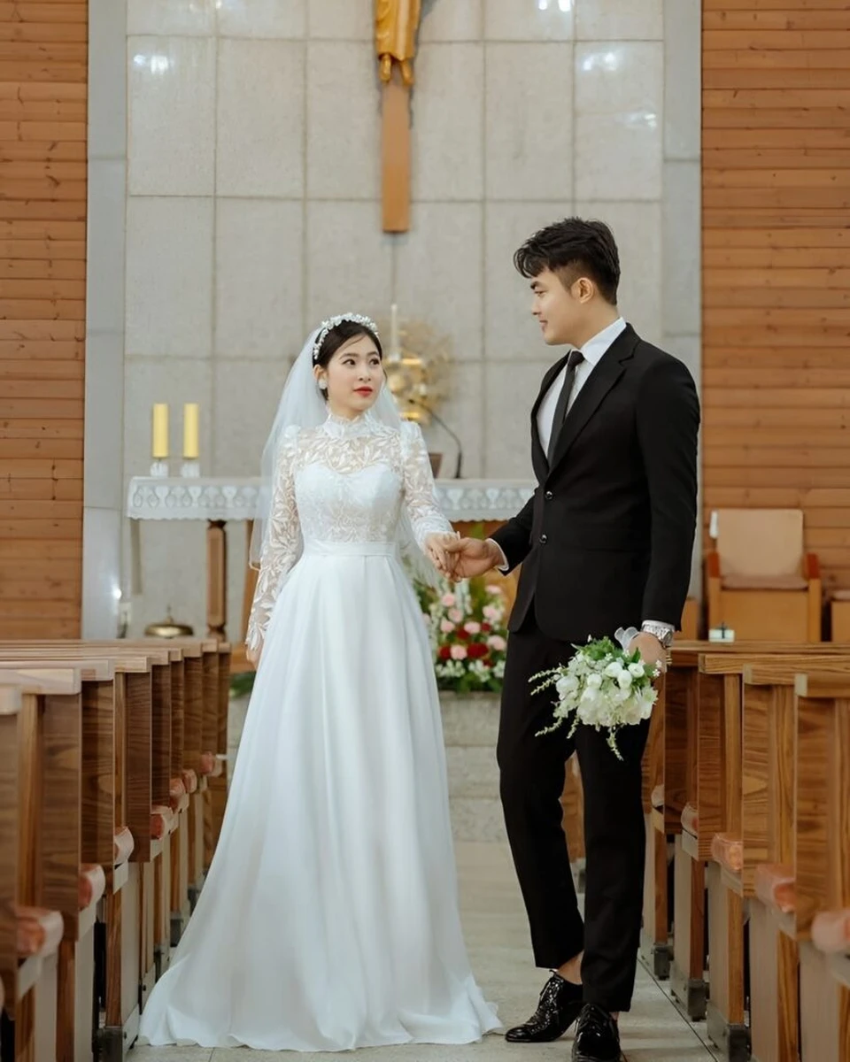 Cô dâu mặc váy cưới trắng ren tay dài kết hợp khăn voan và chú rể diện vest đen cầm hoa cưới trắng
