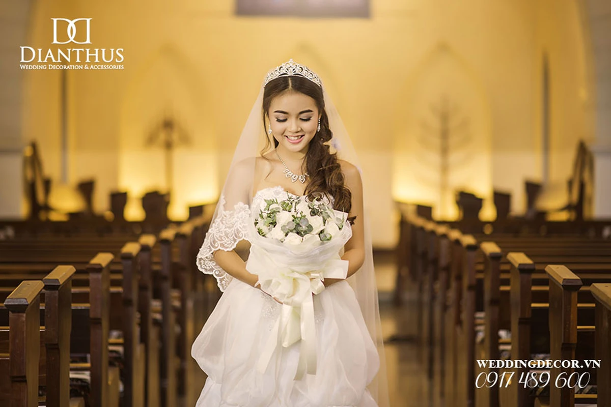 Cô dâu mặc váy cưới trắng cúp ngực kết hợp khăn voan dài và vương miện, cầm bó hoa cưới trắng