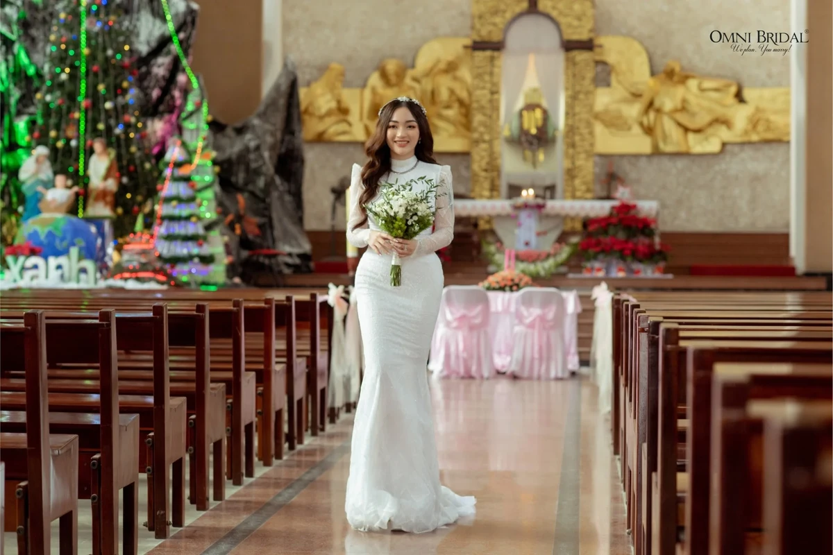 Cô dâu diện váy cưới trắng dáng ôm, tay dài, cổ cao đứng trong không gian thánh đường trang trọng