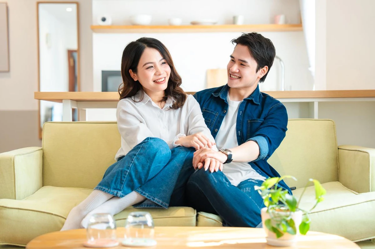 Cặp vợ chồng trẻ mỉm cười hạnh phúc và nắm tay nhau khi ngồi trên ghế sofa phòng khách