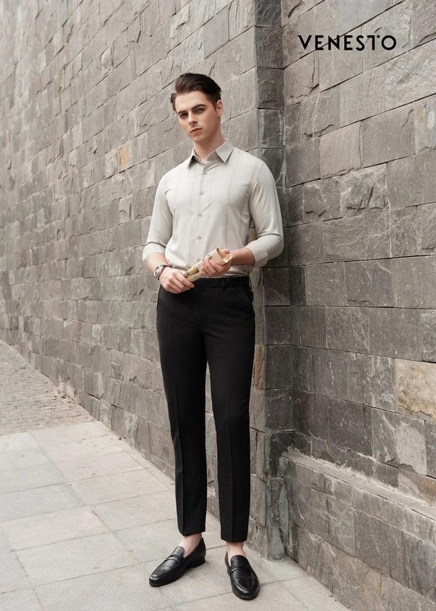 Nam mẫu mặc áo sơ mi màu be tay dài, quần tây đen, đứng tựa tường đá với phong cách thanh lịch