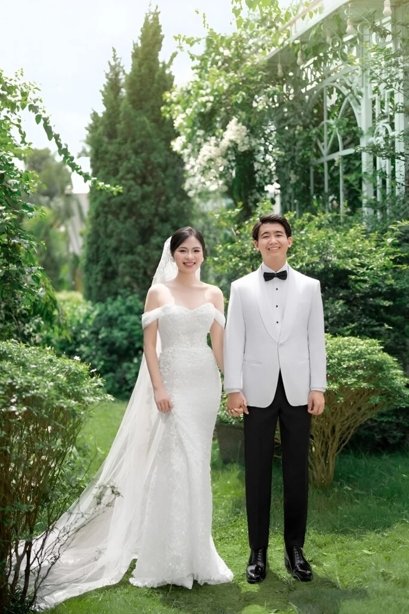 Cặp đôi rạng rỡ nắm tay nhau chụp ảnh cưới tại phim trường sân vườn xanh mát, cô dâu mặc váy cưới trễ vai thanh lịch bên chú rể diện vest trắng